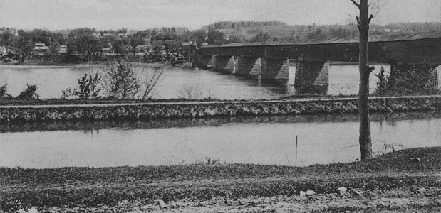 This photograph shows the Delaware and Raritan Feeder Canal in the Borough of Stockton, Hunterdon County, and the Centre Bridge-Stockton covered bridge over the Delaware River.  The covered bridge was destroyed by a fire caused by a lightning strike in 1923.  The current steel truss bridge over the Delaware was constructed in 1927 and rests upon the stone piers that once supported the wooden covered span.  Note the relative absence of vegetation along the canal towpath.  Although the Delaware and Raritan Canal State embankment is heavily-vegetated today, during the canal's period of operation this was not the case.  The Canal Company ensured that the area near the towpath was kept free of trees and plants, which could interfere with boat masts, sails and towlines. 